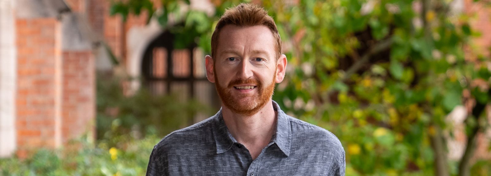 Portrait of a ginger haired and bearded smiling man in a light blur shirt, standing outdoors on a university campus, smiling at the camera, with brick buildings and greenery in the background.